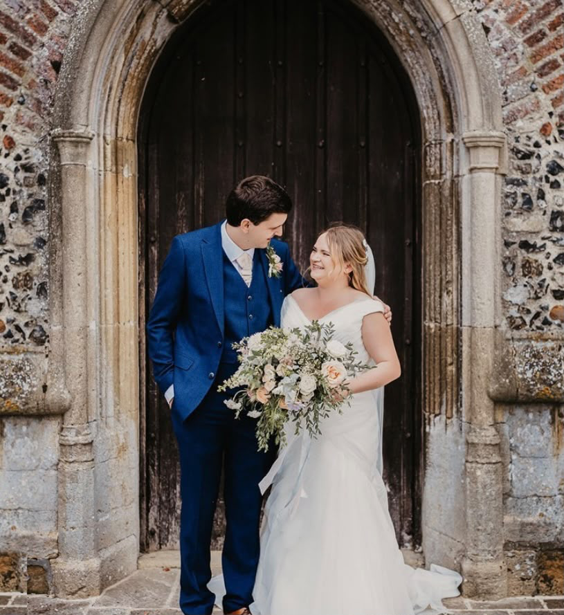 Bride and groom smiling together outside an arched wooden door with a wild ivory and peach bouquet