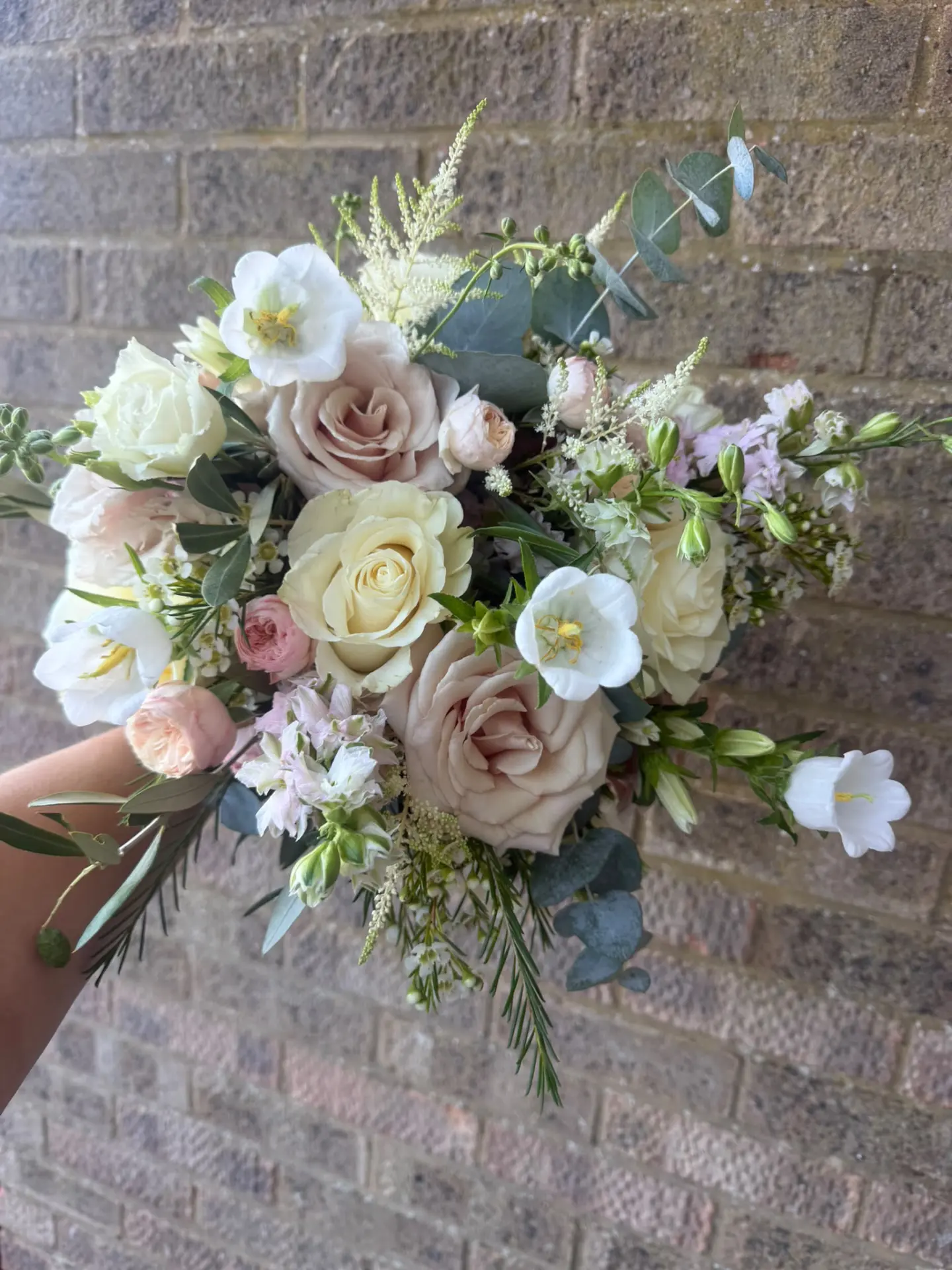 Hand-held bouquet of ivory and dusty mauve roses with sage greenery against a brick wall