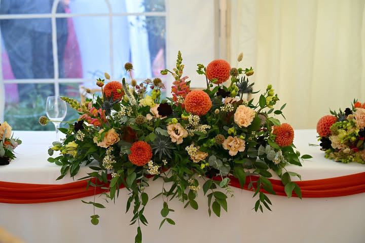 Table centrepiece of orange dahlias, peach carnations and thistles on a dressed marquee table
