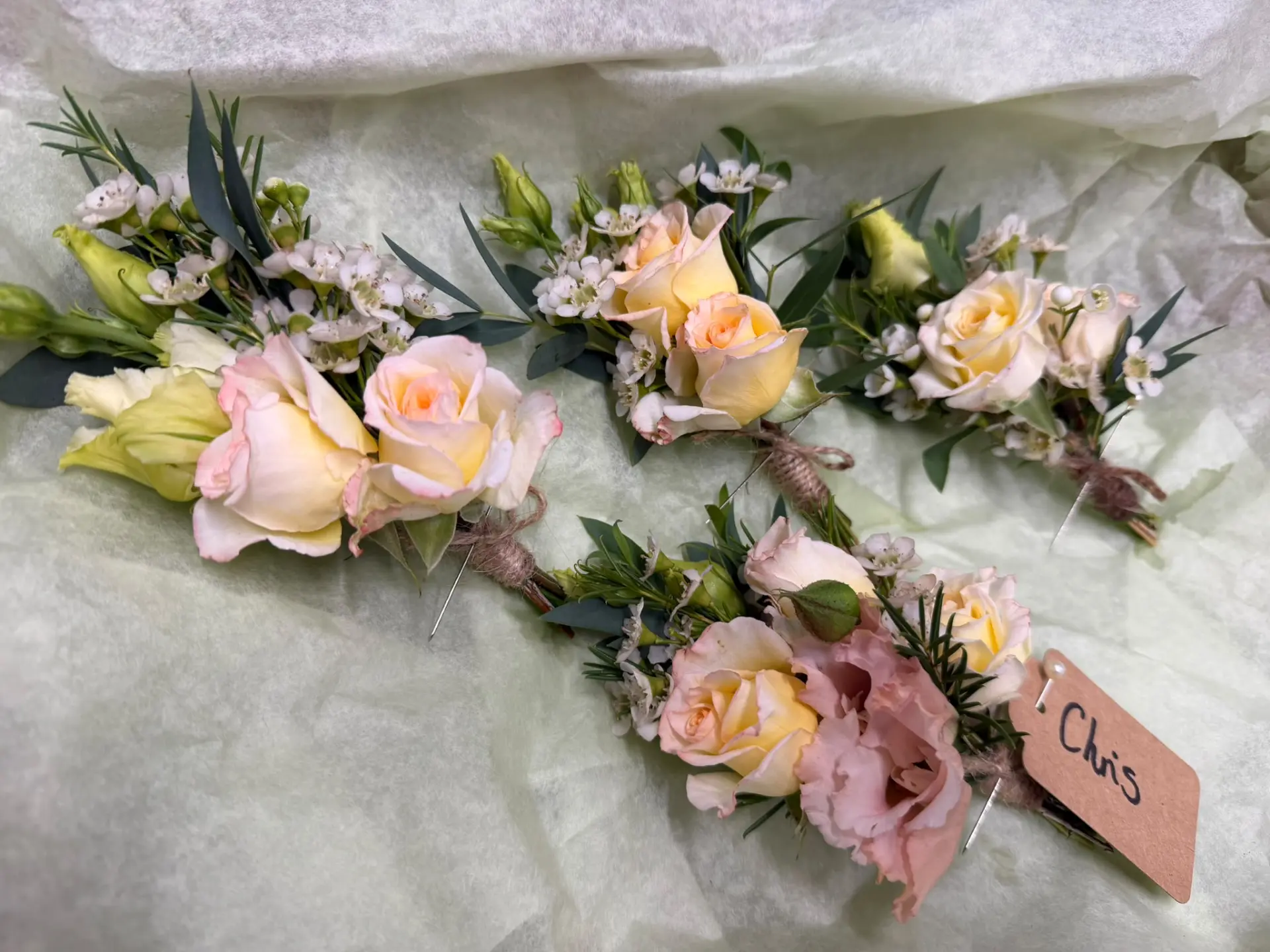 Four floral buttonholes with peach and pink rosebuds resting on tissue paper