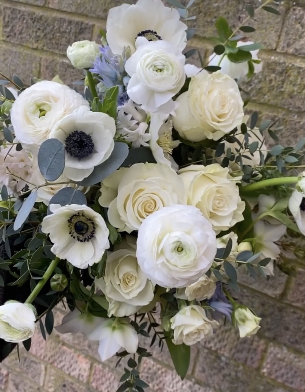 Bridal bouquet of white ranunculus, anemones and cream roses with eucalyptus against a brick wall