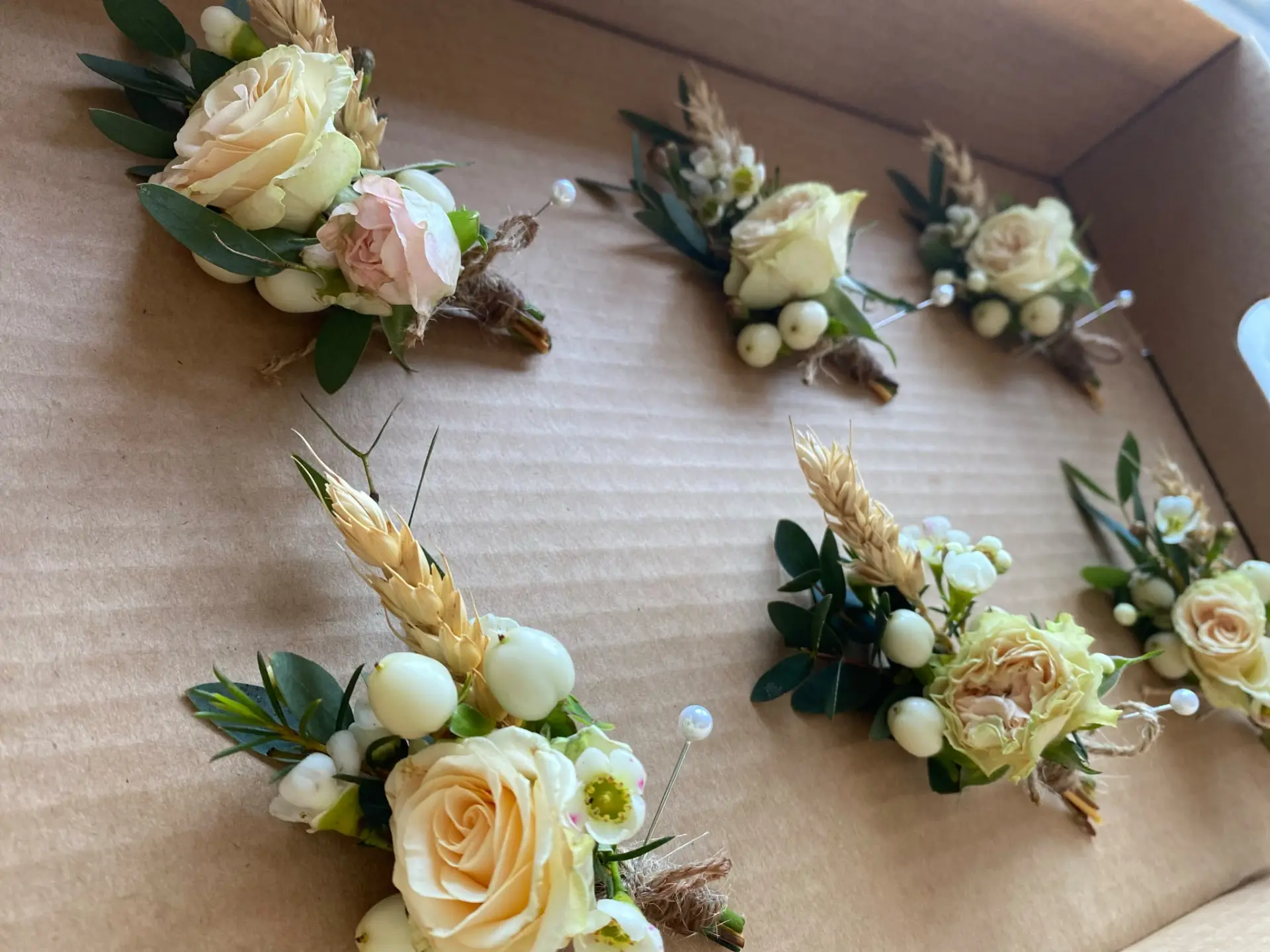 Six buttonholes with cream roses and dried wheat laid in a delivery box