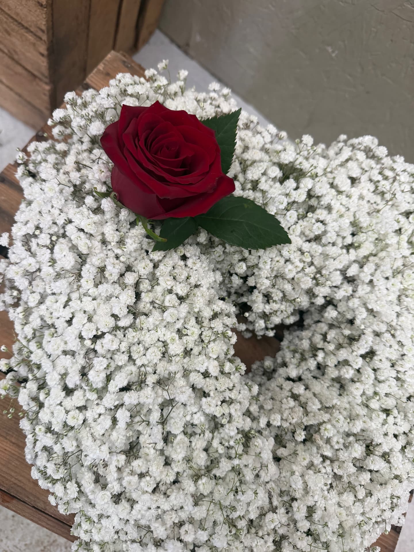 Heart-shaped tribute wreath of gypsophila with a single red rose resting on a wooden crate