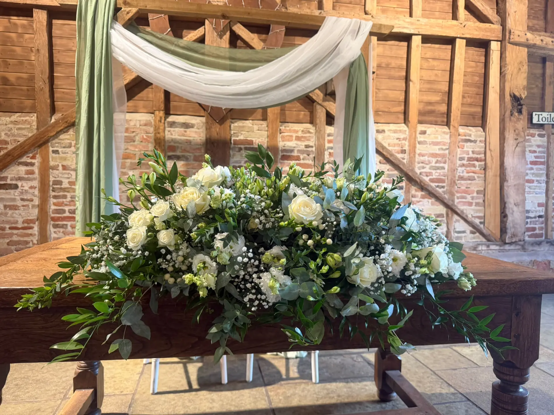 Long low table arrangement of cream roses and greenery on a rustic wooden ceremony table
