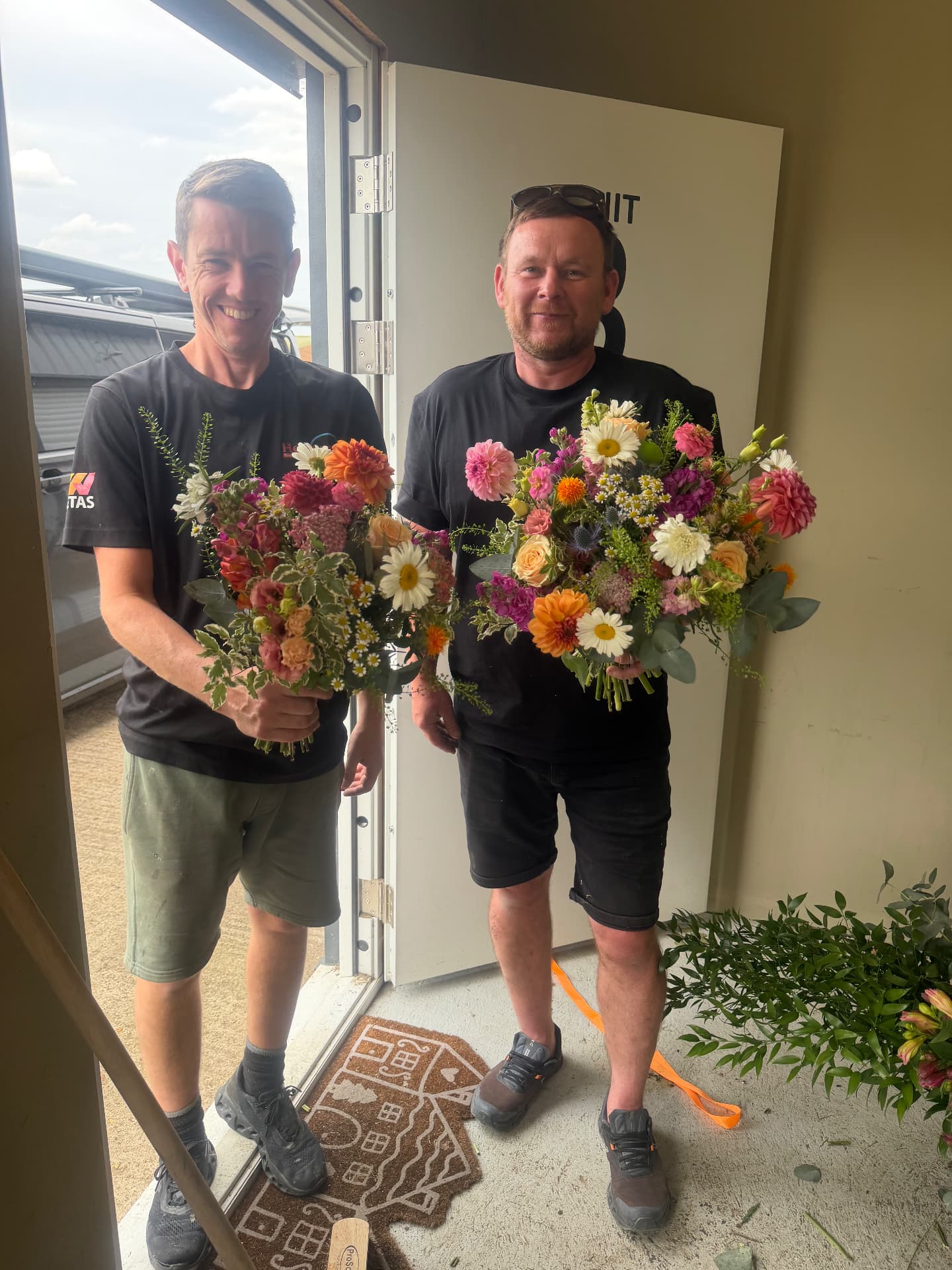 Two men each holding a bouquet, standing in a doorway